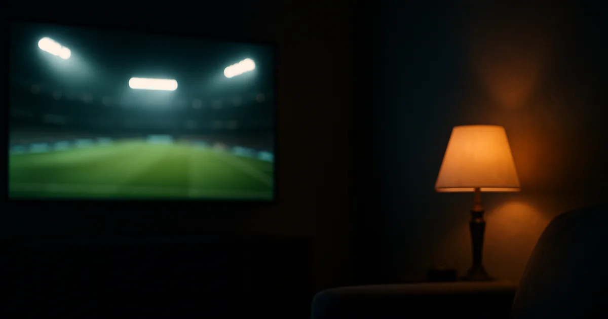 Salon sombre le soir, un grand téléviseur allumé diffusant un match de football sur une pelouse éclairée par les projecteurs d'un stade, canapé vide et télécommande posée sur l'accoudoir.